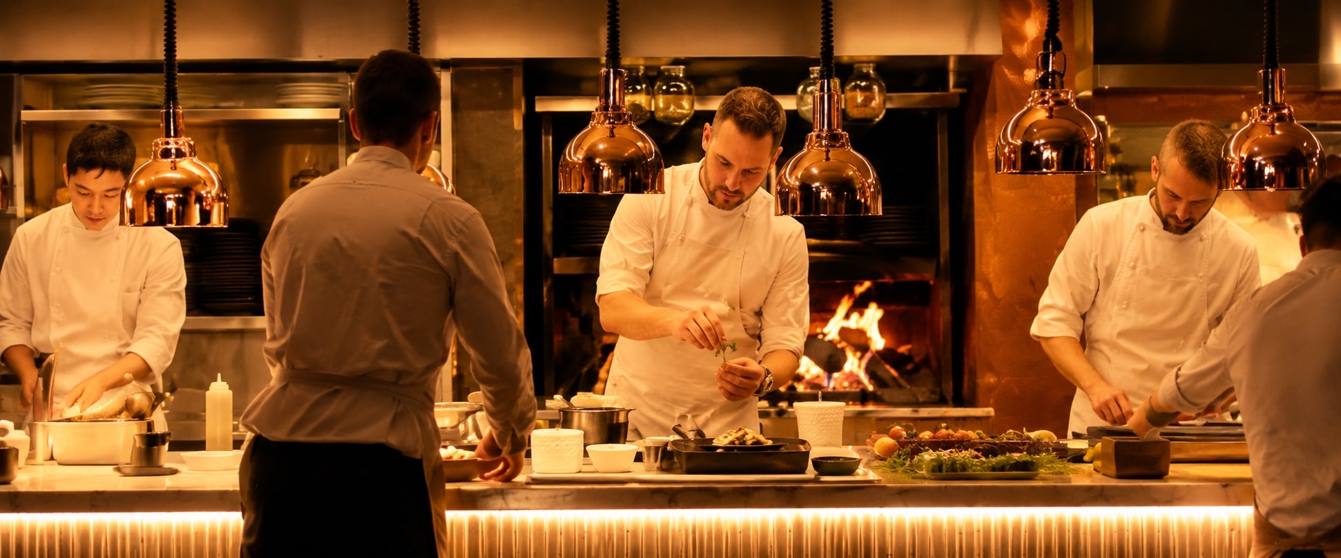 Cooks working an open kitchen pass under copper lamps