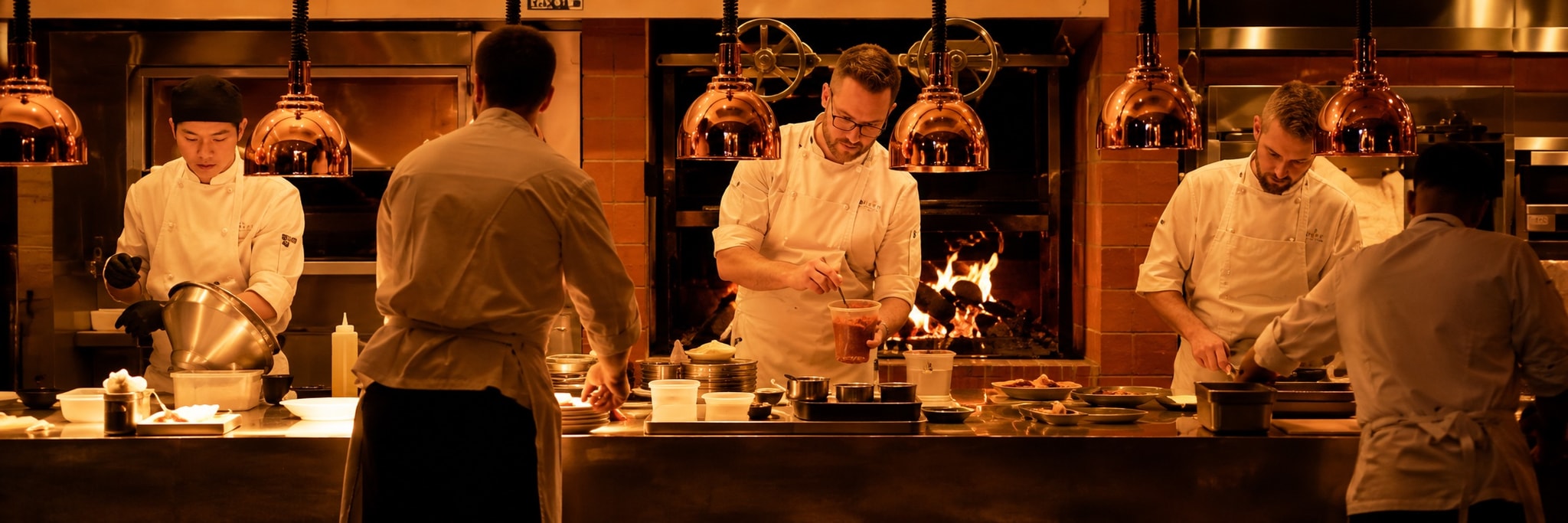 Chefs working an open kitchen pass beside a wood fire