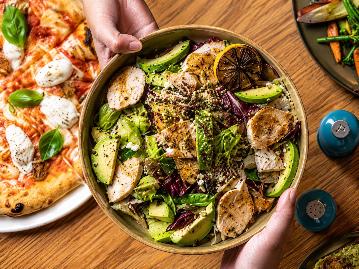 Hands setting down a chicken and avocado bowl beside a pizza